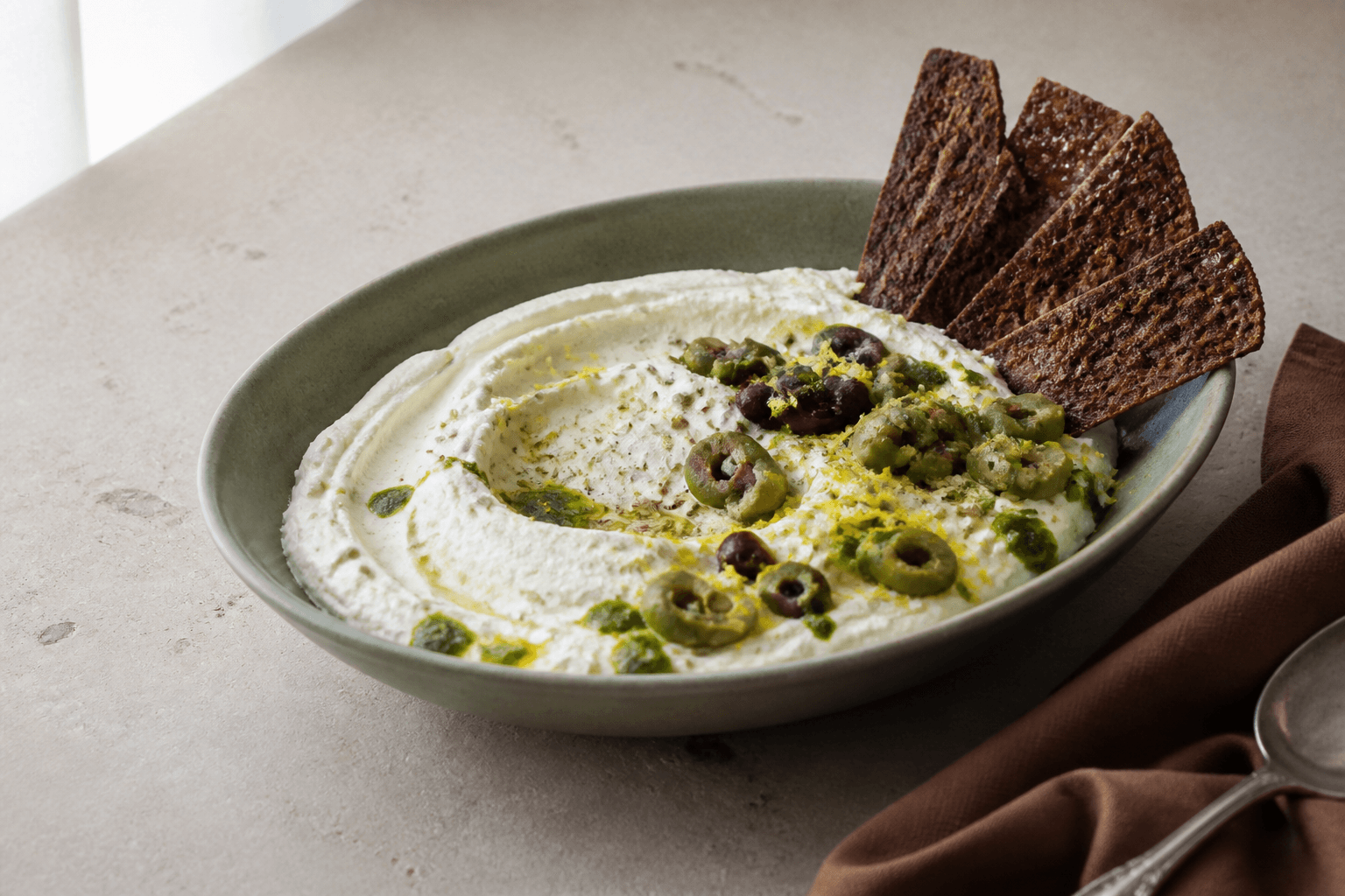 Pisket ricottadip med grønne og mørke oliven, citron, urteolie og sprøde rugflager i salviegrøn skål