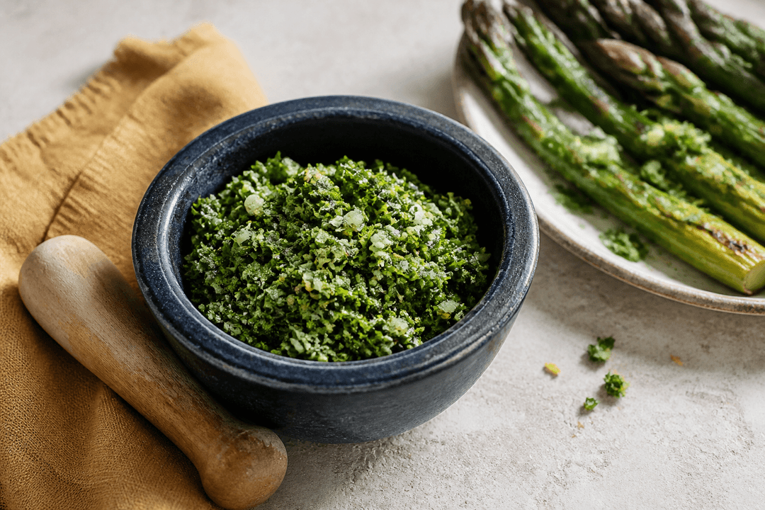 Grov grøn gremolata med persille, citronskal, hvidløg, kapers og pistacie i lille mørkeblå morter ved grillede asparges