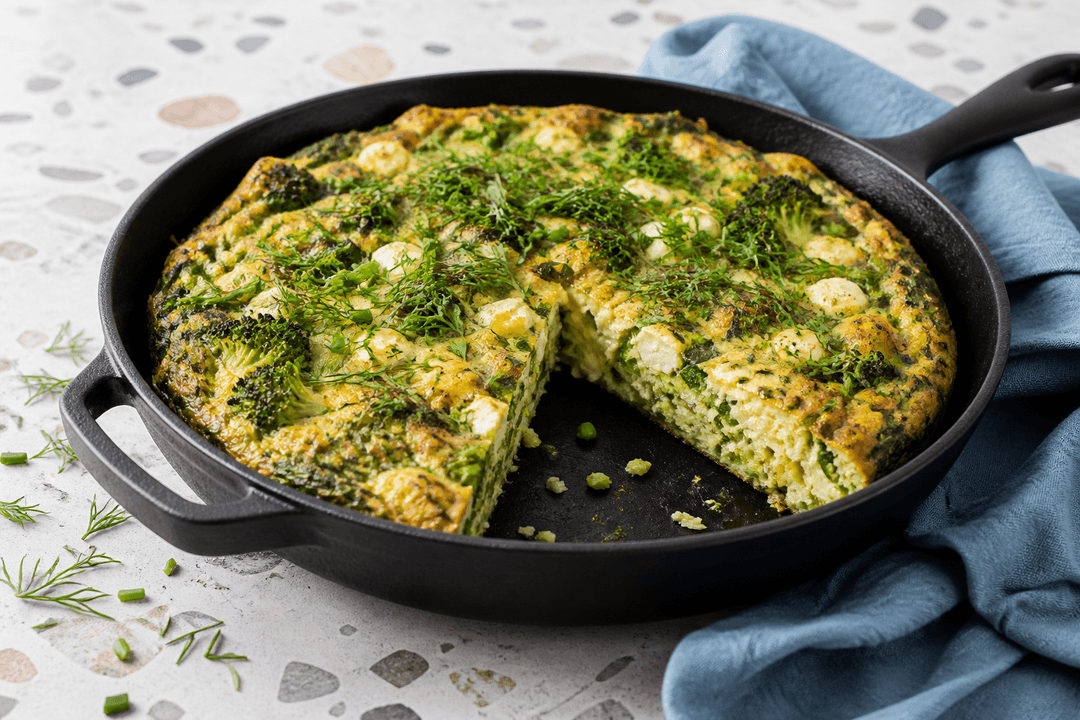 Tyk ovnbagt æggekage i sort støbejernspande med broccoli, porre, spinat, ærter, gedeost, dild og purløg samt ét udskåret stykke