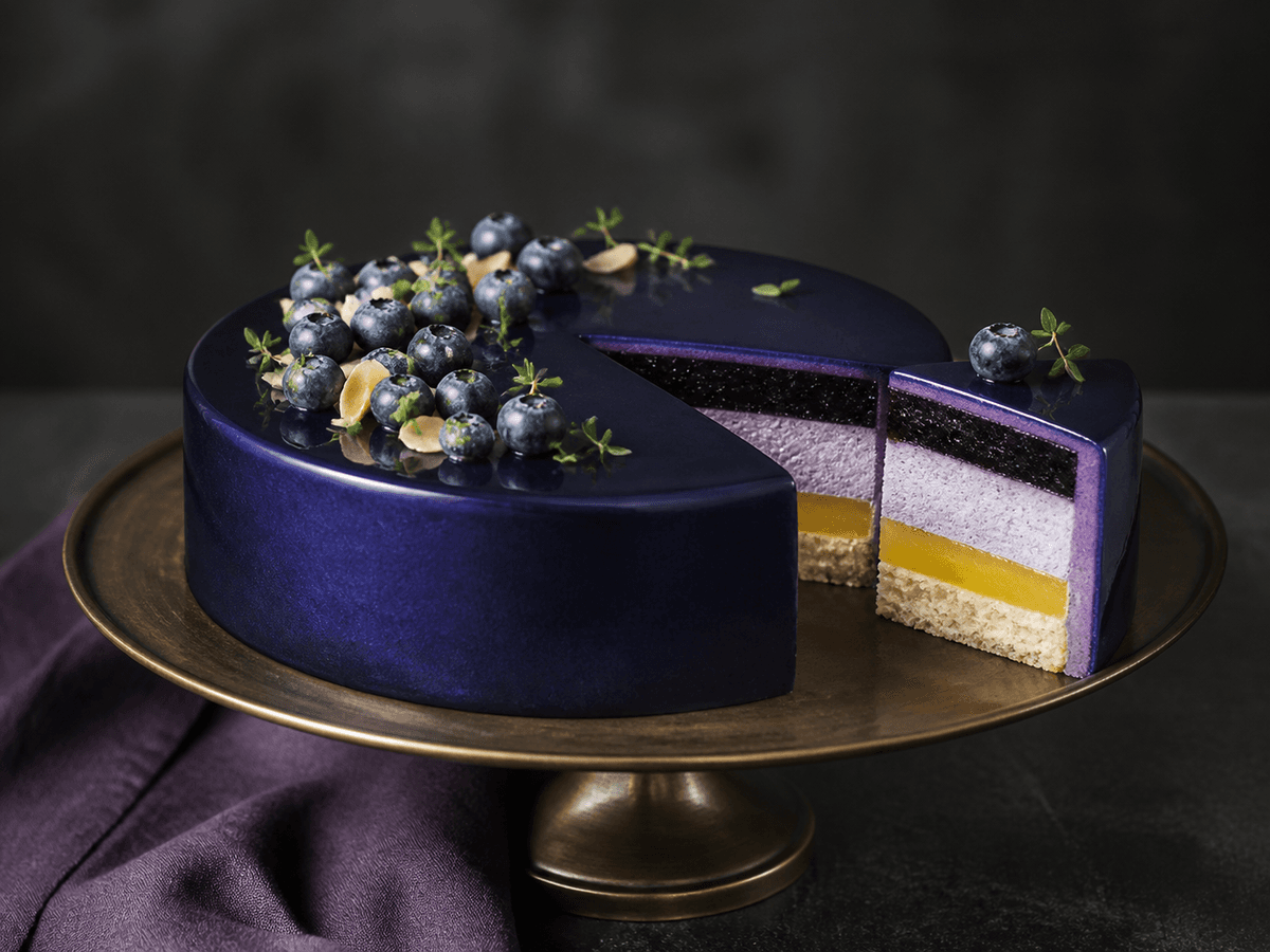 Rund blåbærmoussekage med mørk midnatsblå spejlglaze, blåbær og mandelflager, udskåret så mandelbund, citronkerne, mousse og blåbærgel ses