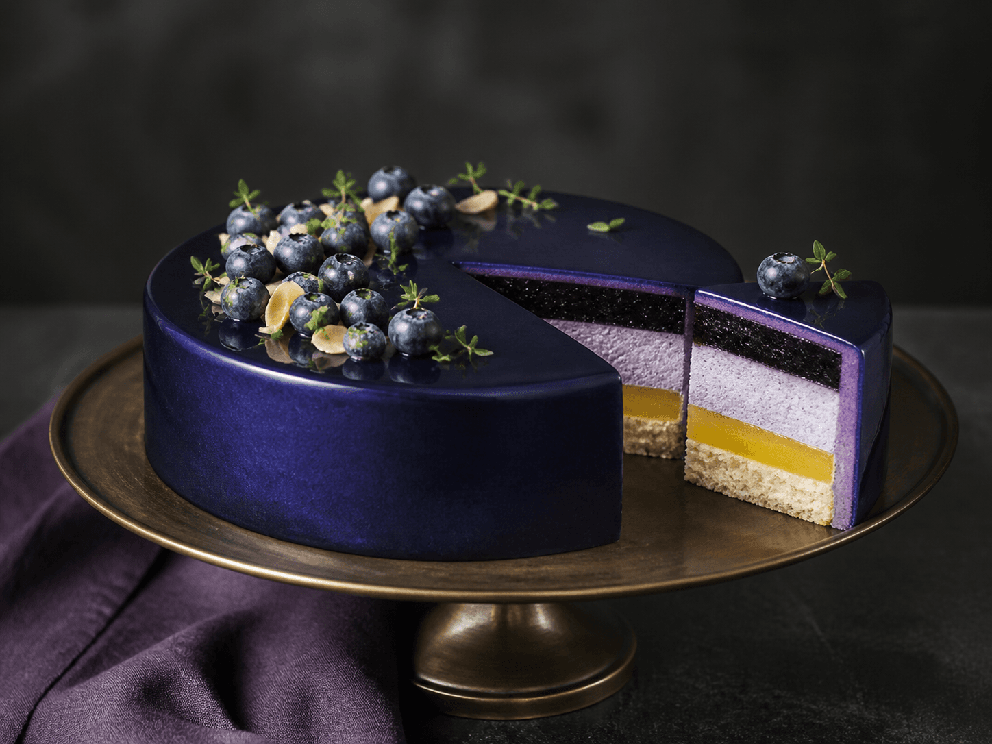 Rund blåbærmoussekage med mørk midnatsblå spejlglaze, blåbær og mandelflager, udskåret så mandelbund, citronkerne, mousse og blåbærgel ses