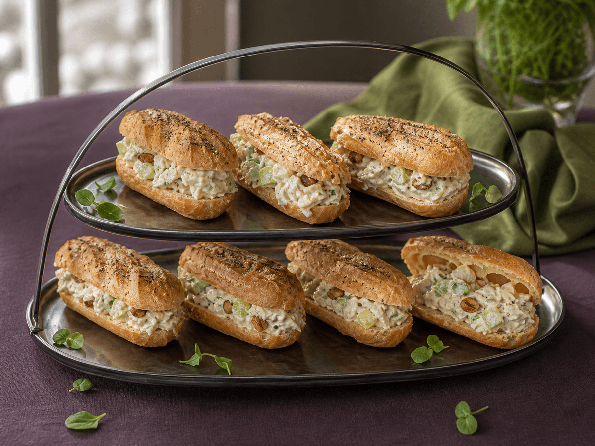 Otte små aflange vandbakkelses-eclairs fyldt med cremet kyllingesalat, grønne æbletern, selleri, hasselnød og estragon på mørkt sølvfad