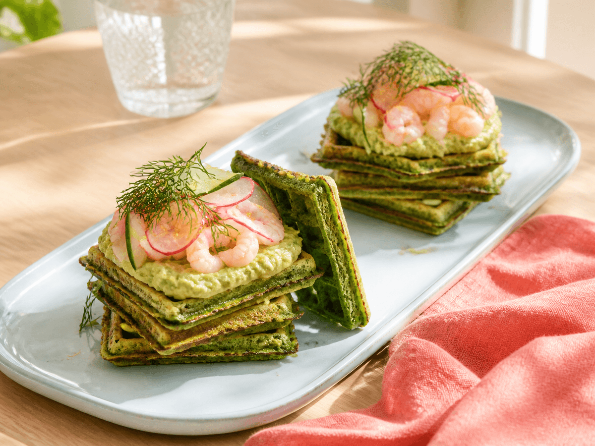 To stabler firkantede grønne spinatvafler med avocado-ærtecreme, rejer, radise, agurk, dild og citron på lyseblåt fad