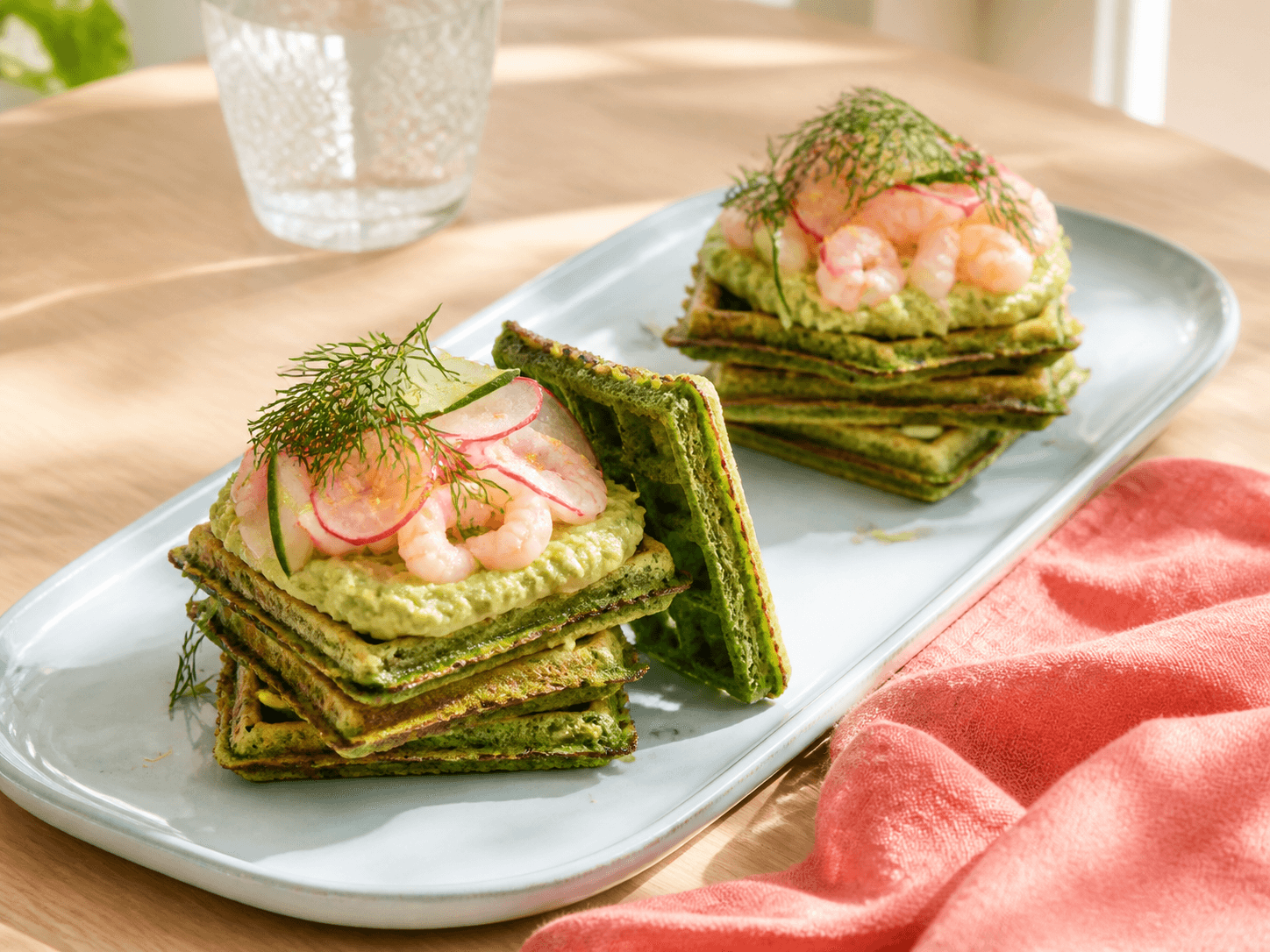 To stabler firkantede grønne spinatvafler med avocado-ærtecreme, rejer, radise, agurk, dild og citron på lyseblåt fad