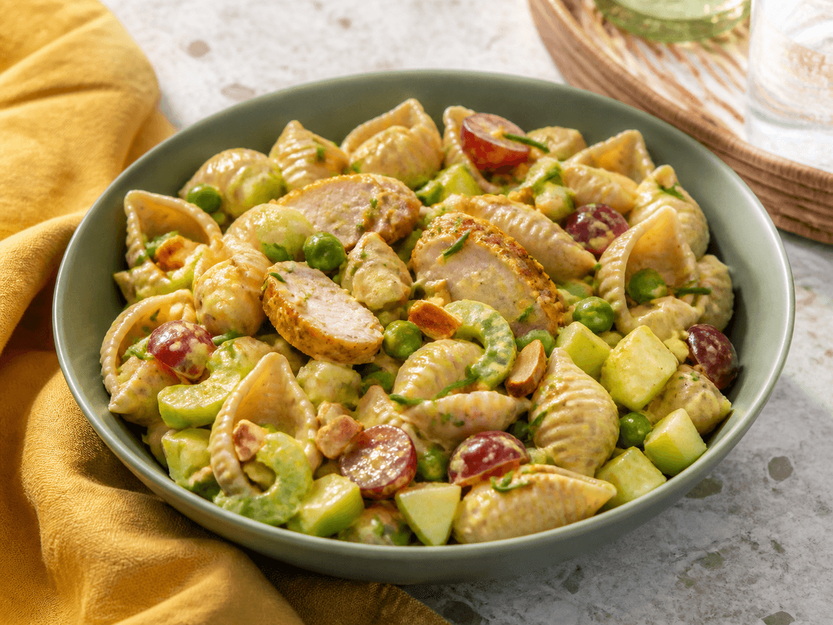 Kold fuldkornsskalpastasalat med gylden kylling, æble, selleri, ærter, røde druer, mandler og let mango-karrydressing i salviegrøn skål