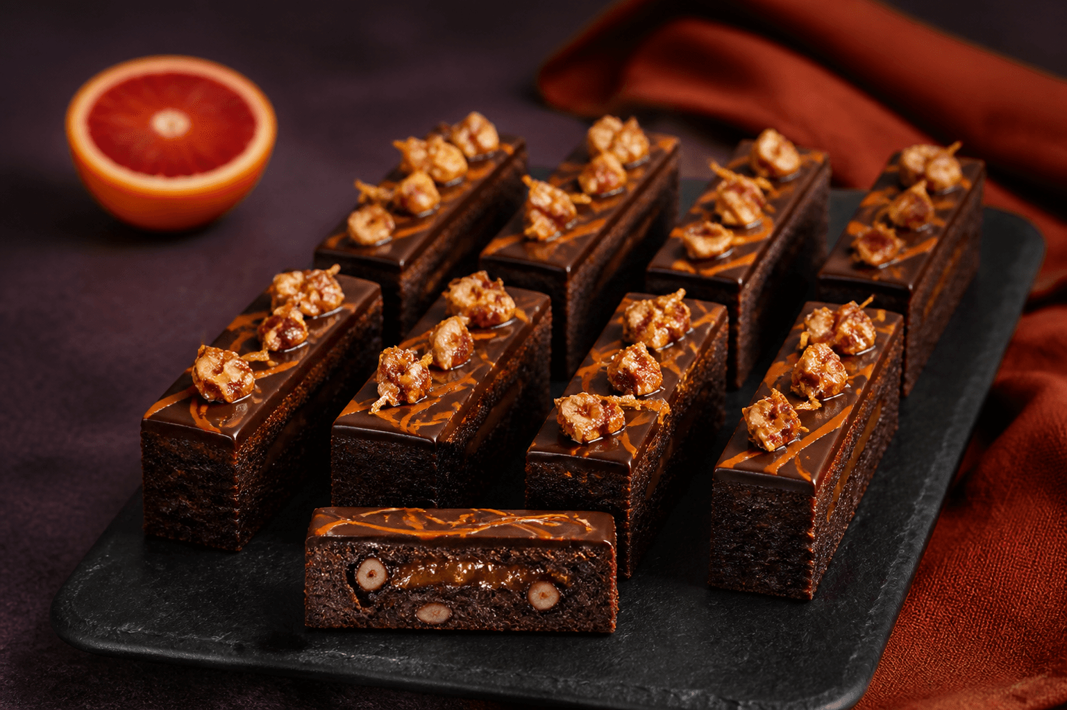 Ni høje rektangulære blodappelsinbrownies med mørk ganache, orange marmormønster, hasselnøddepraline og ét åbent stykke på sort skifer