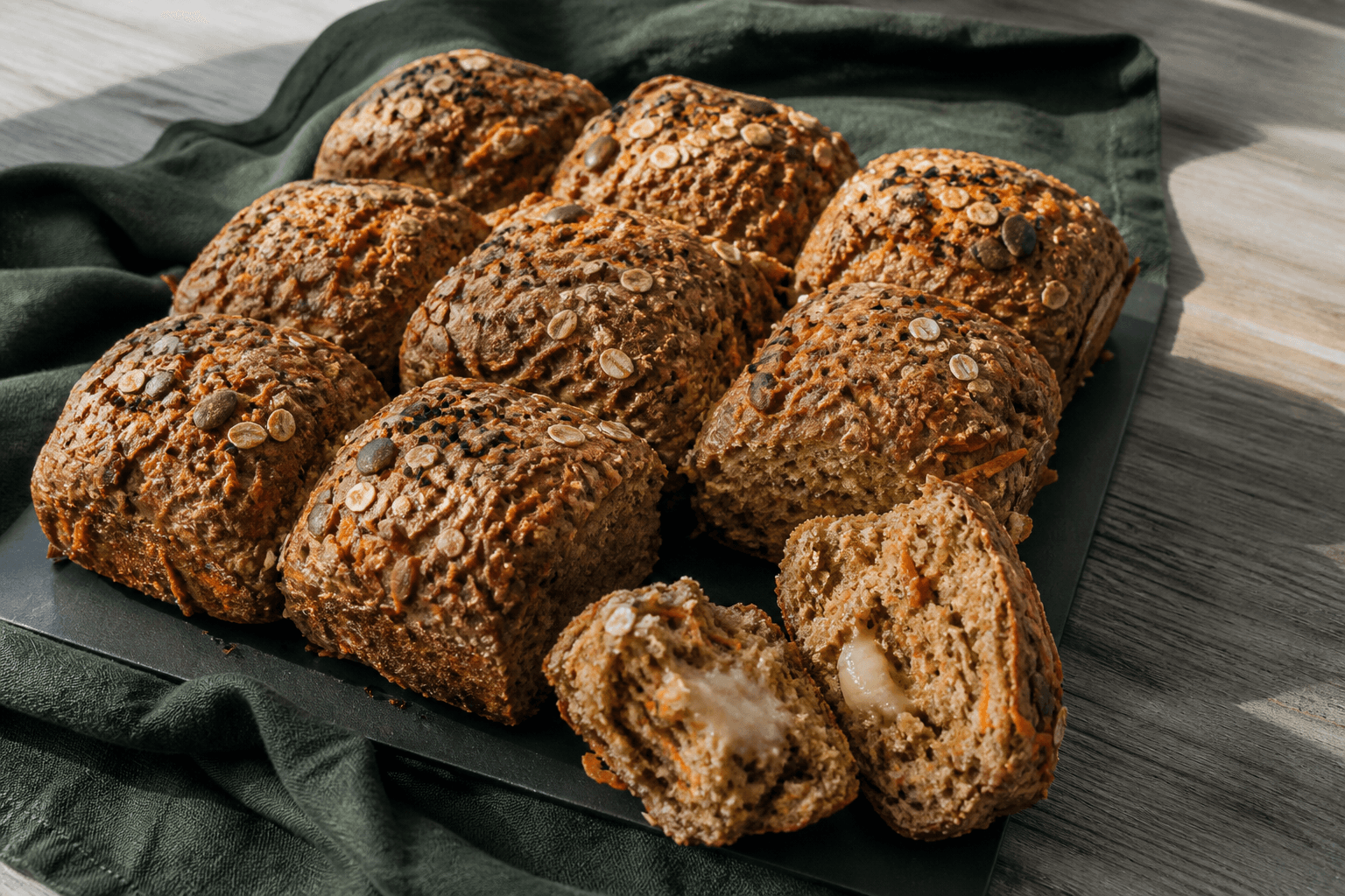 Otte mørkgyldne rektangulære gulerods-rugbrud med rugflager, græskarkerner og nigella på sort bagestål, ét delt med smeltet ostekerne