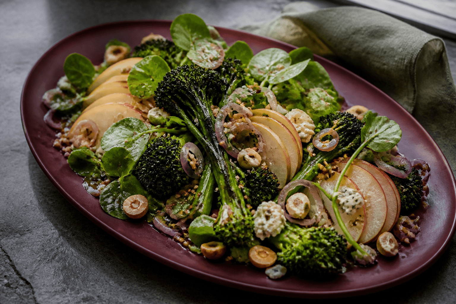 Burgunderfarvet ovalt fad med grillet broccoli, røde pærevifter, blå ost, ristet boghvede, syltede skalotteløg, brøndkarse og hasselnød