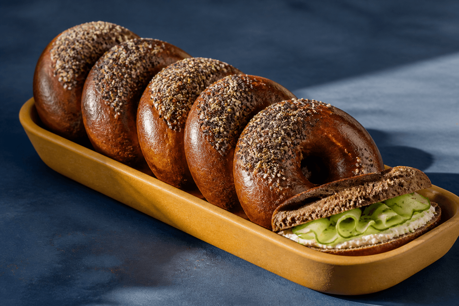 Seks blanke mørke rugmalt-bagels med halv frøtop i sennepsgult fad, én delt med peberrodscreme og agurk