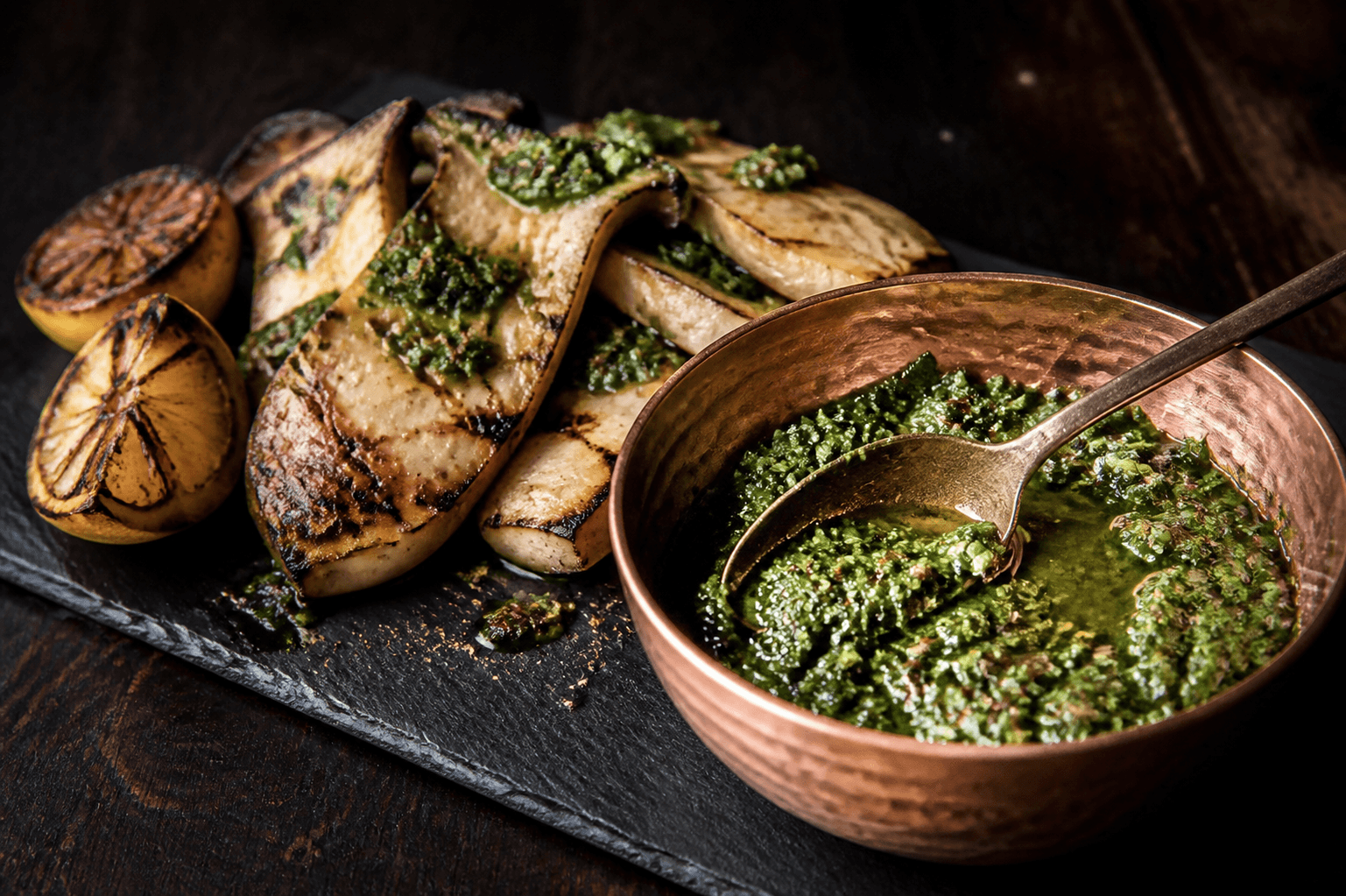 Grov mørkegrøn chimichurri med grillet forårsløg, persille, mynte, oregano, syltet citron, chile og korianderfrø i hamret kobberskål ved grillede kejserhatte