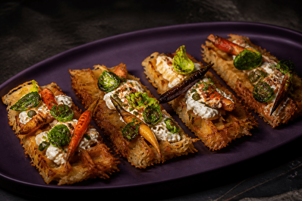 Fire lange sprøde kartoffel-selleri-röstivafler med ristede farvede gulerødder, blistrede rosenkålsblade, pisket røget mozzarella og kapers-persilleolie på auberginefad