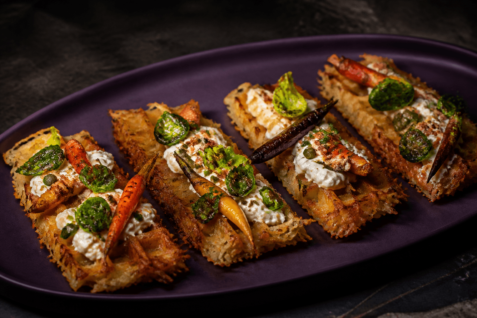 Fire lange sprøde kartoffel-selleri-röstivafler med ristede farvede gulerødder, blistrede rosenkålsblade, pisket røget mozzarella og kapers-persilleolie på auberginefad