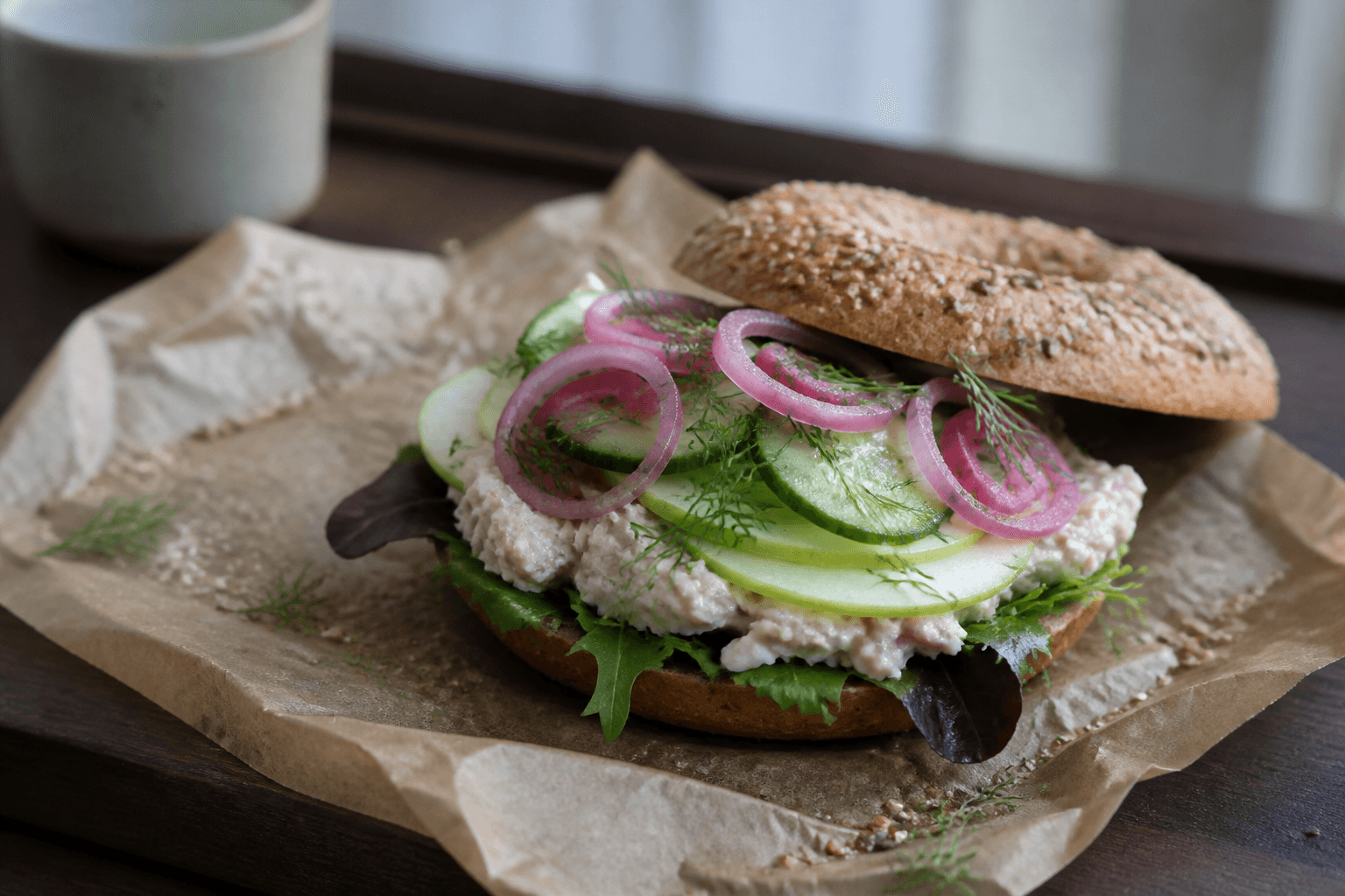 Halvåben ristet rugbagel med grov peberrodsskinkesalat, grønne æbleskiver, dildagurk, salat og syltet rødløg på pergament