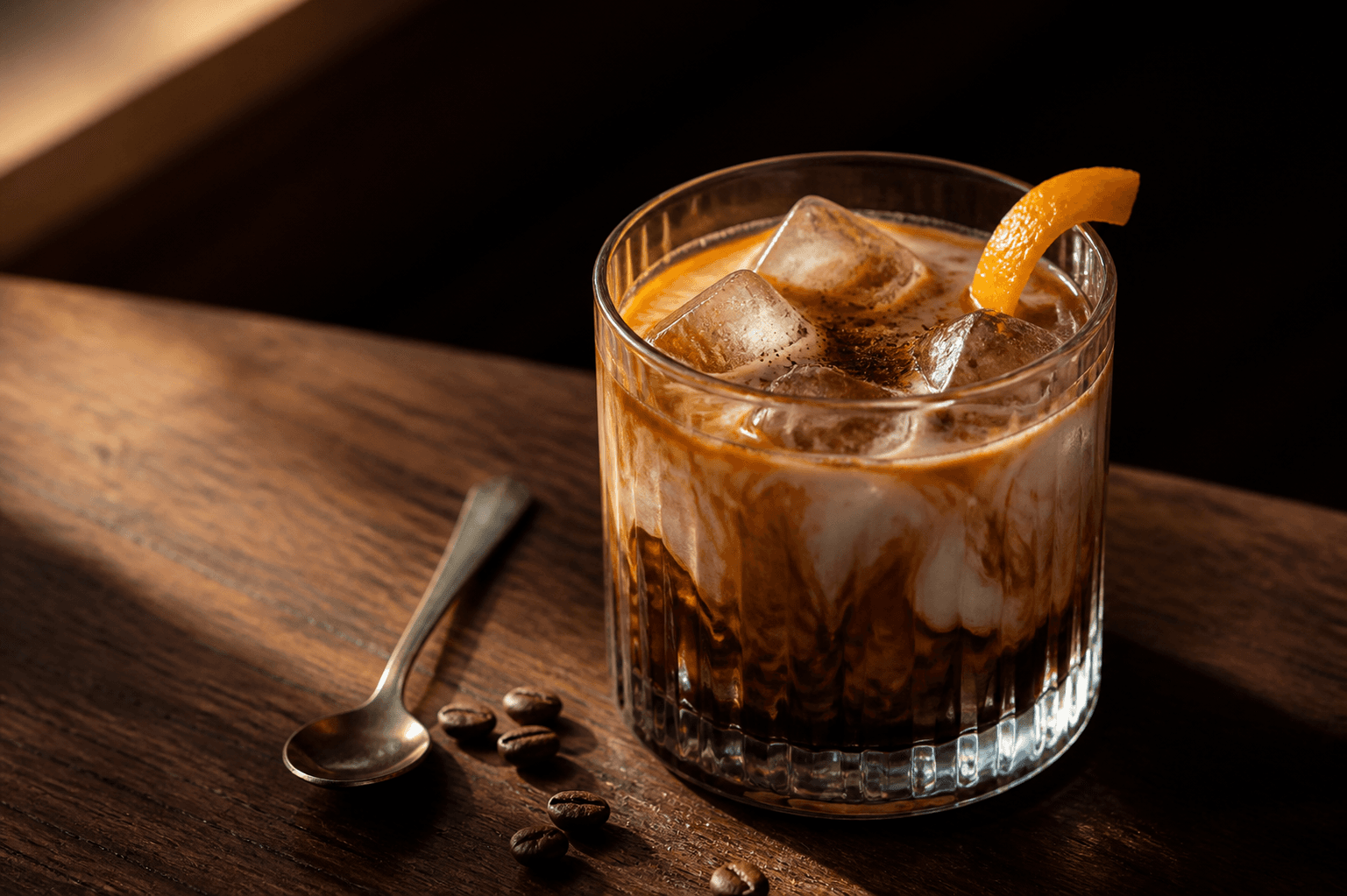 Lavt riflet glas med is, mørk kardemomme-koldbryg, hvirvlet havrefløde, whiskykaramel og appelsinskal på mørkt træ