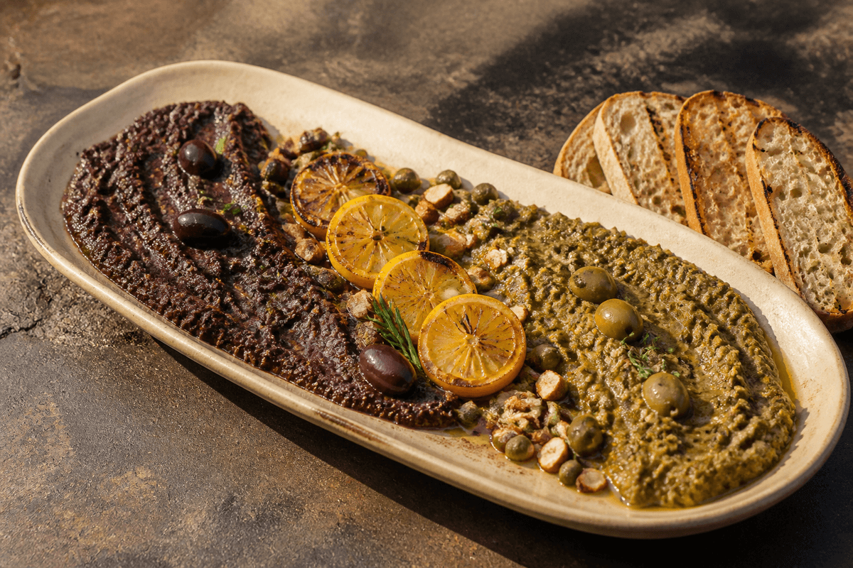 Langt sandfarvet fad med separate grove sorte og grønne oliventapenader, bagte citronskiver, mandler, kapers, rosmarin og grillede brød
