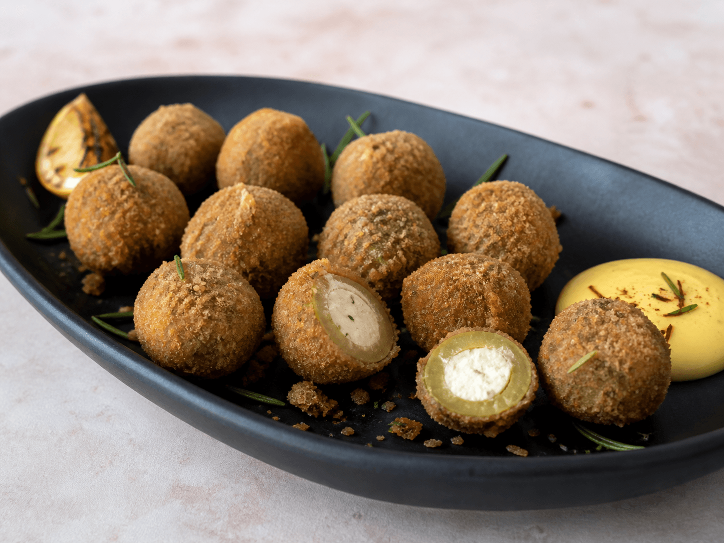 Langt sort fad med rugpanerede friterede grønne oliven, to åbne med citron-gedeostkerne, safranaioli, rosmarin og grillet citron