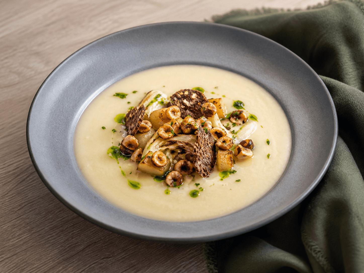 Bred grå skål med silkeblød lys hvidkålssuppe, karamelliseret kål, ristede pæretern, brunede hasselnødder, grøn persilleolie og mørke rugchips
