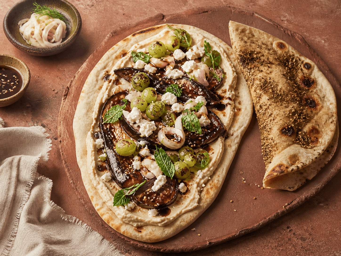 Åbent ovalt zaatarfladbrød med butterbean-hummus, blanke harissa-auberginebåde, grillede grønne druer, syltet fennikel, feta, mynte og sesam