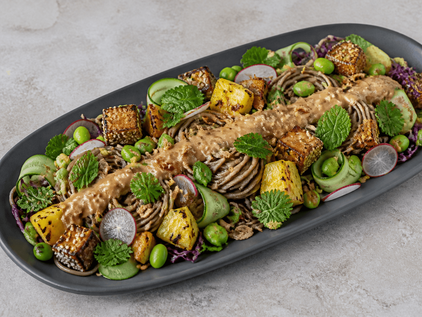 Langt mørkt fad med kolde sobanudler, sprød sesamtofu, grillet ananas, agurk, rødkål, edamame, radise, urter og et centralt bånd peanutdressing