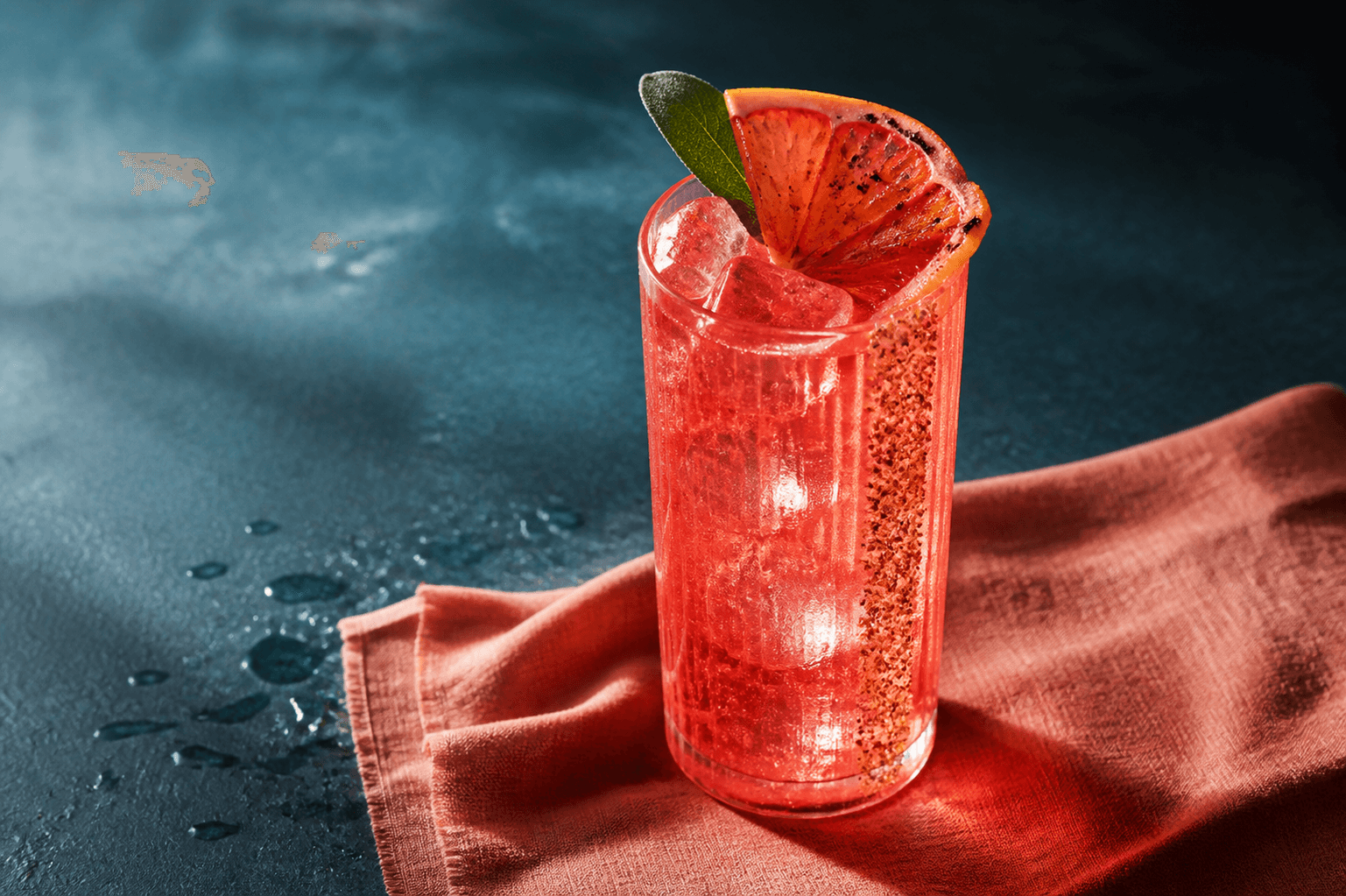 Høj rillet highball med rubinrød boblende Paloma, klar spydis, grillet grapefrugt, salvie og lodret stribe røget grapefrugtsalt