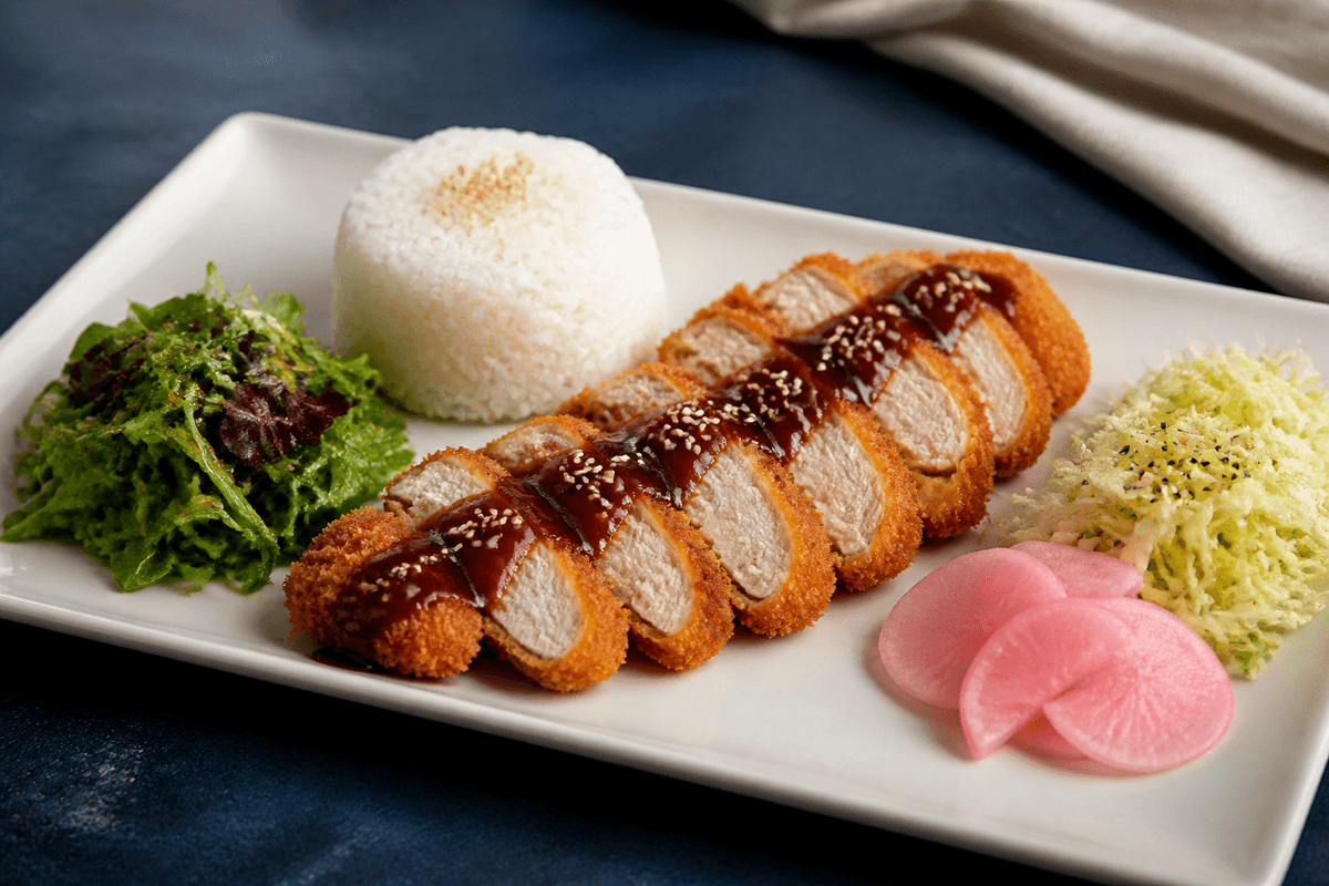 Lang elfenbensfarvet tallerken med gennemstegt sprød kylling-katsu, mørk æble-misosauce, ris, sesamkål, grøn salat og syltet radise
