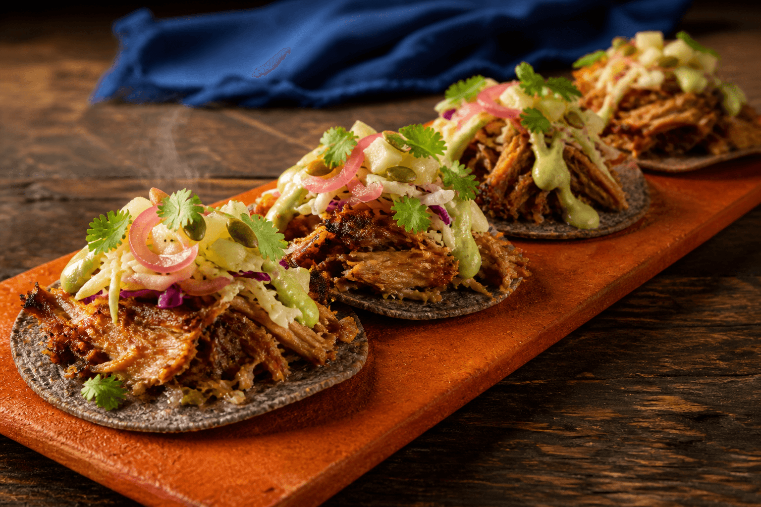 Langt brændt orange fad med fire sprøde blå majstostadas, gennemtilberedt appelsin-carnitas, ananas-jicamaslaw, avocadocreme, syltet rødløg og græskarkerner