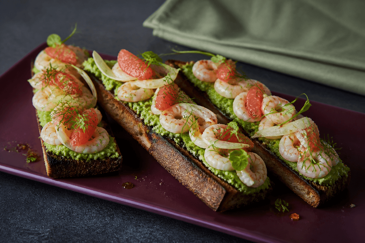 Langt auberginefarvet fad med fire grillede surdejsbrød, avocado-ærtemos, fuldt tilberedte citronrejer, pink grapefrugt, syltet fennikel og dild