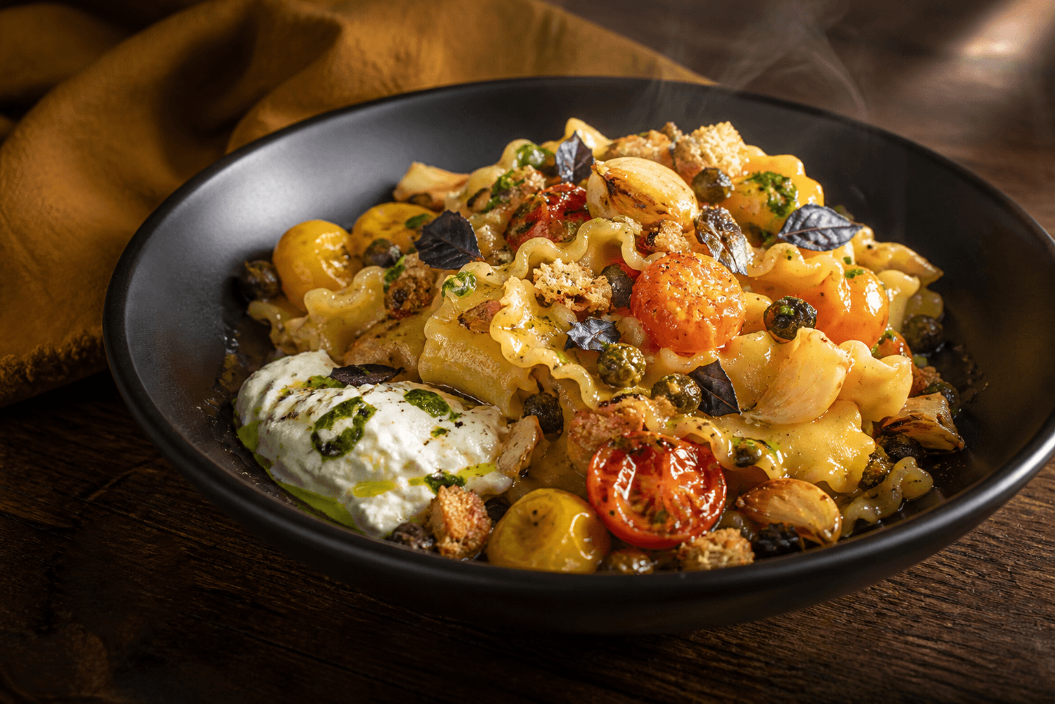 Mat sort skål med dampende mafaldine, blisterede gule og orange tomater, varme hvidløgsconfitfed, stracciatella, sprøde kapers, lilla basilikum og surdejscrunch