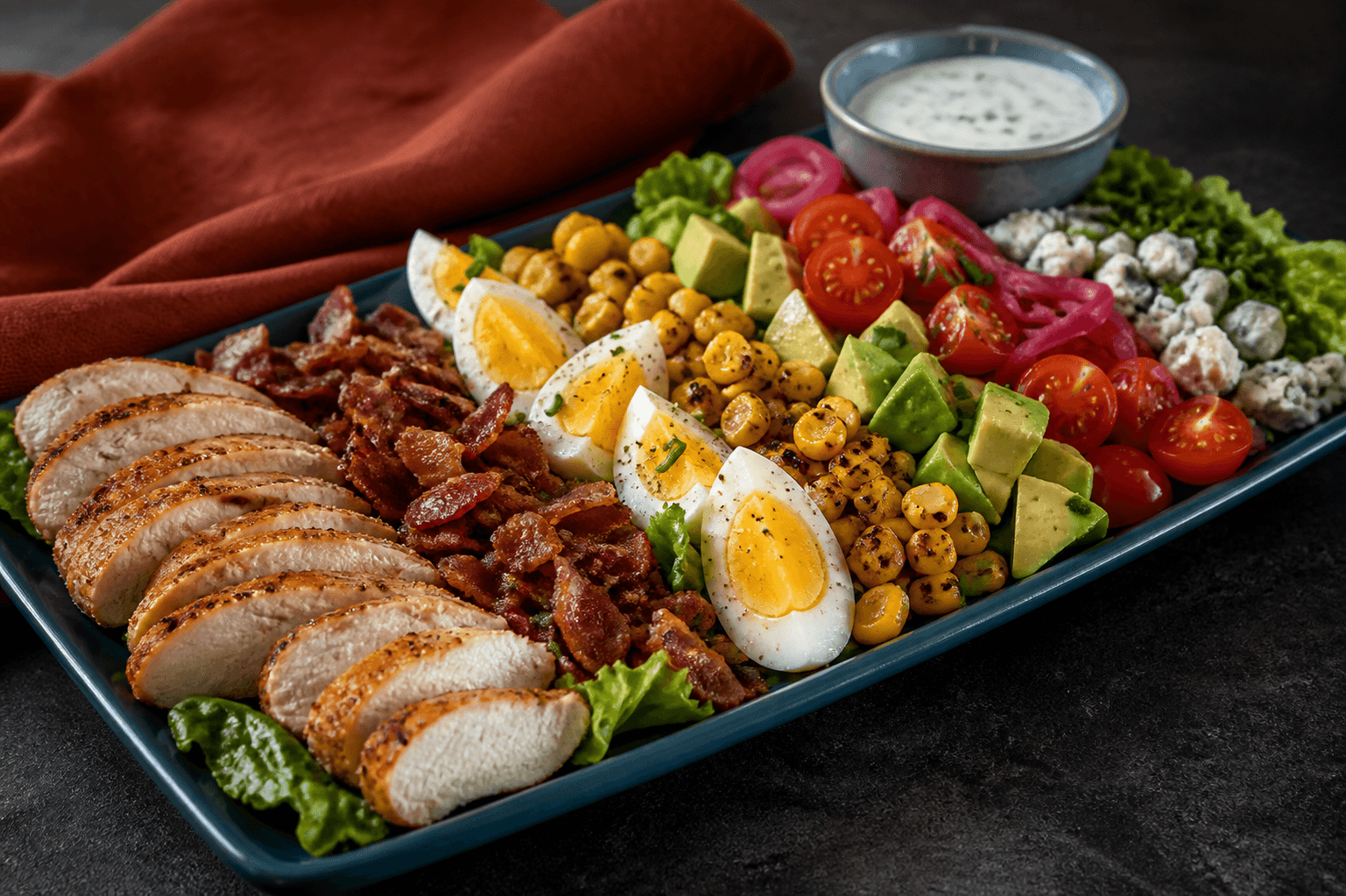 Petrolblåt langt fad med skiver af gennemstegt paprikakylling, sprød bacon, æg, grillet majs, avocado, tomat, syltet rødløg, blåskimmelost og kærnemælksdressing