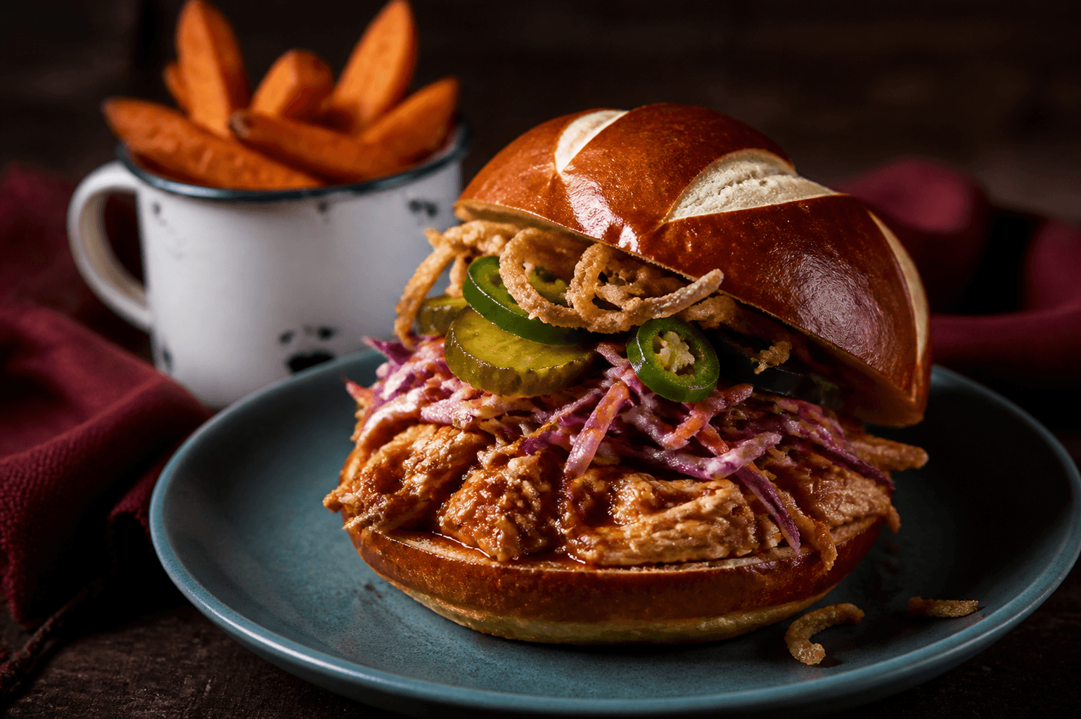 Blank pretzelbrioche-burger med gennemstegt chipotle-pulled chicken, sprød æble-rødkål, pickles, jalapeño og ristede løg på teal tallerken