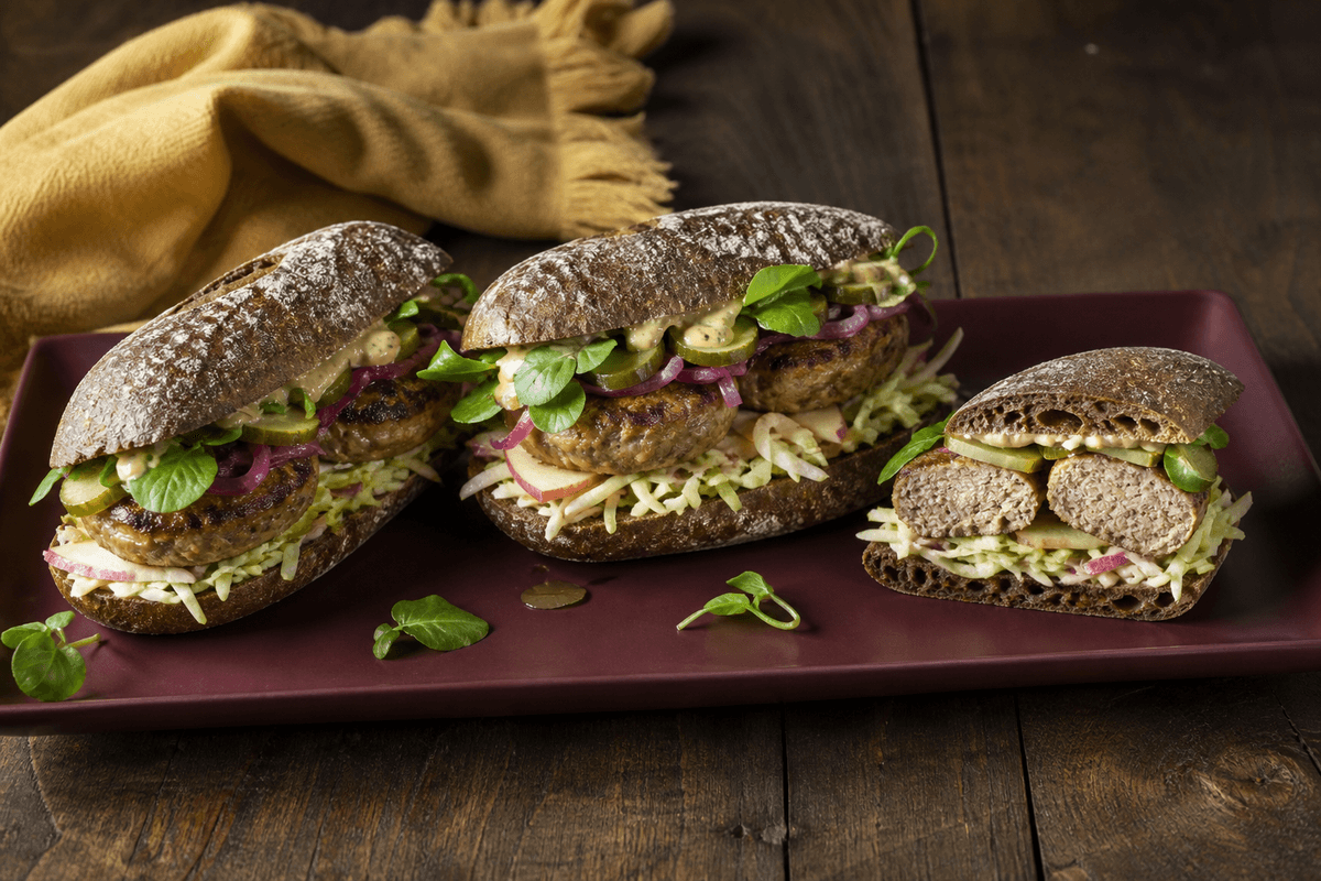 Tre mørke surdejssandwich med fuldt gennemstegte skiver af kalv-flæsk-frikadeller, æble-kålslaw, syltet rødløg, pickles og sennepsremoulade