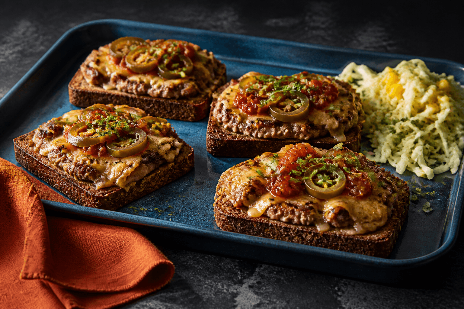 Petrolblå bageplade med fire åbne mørke cowboytoasts med fuldt gennemstegt hakket okse, løg, cheddar, jalapeño og tomatrelish samt kålsalat