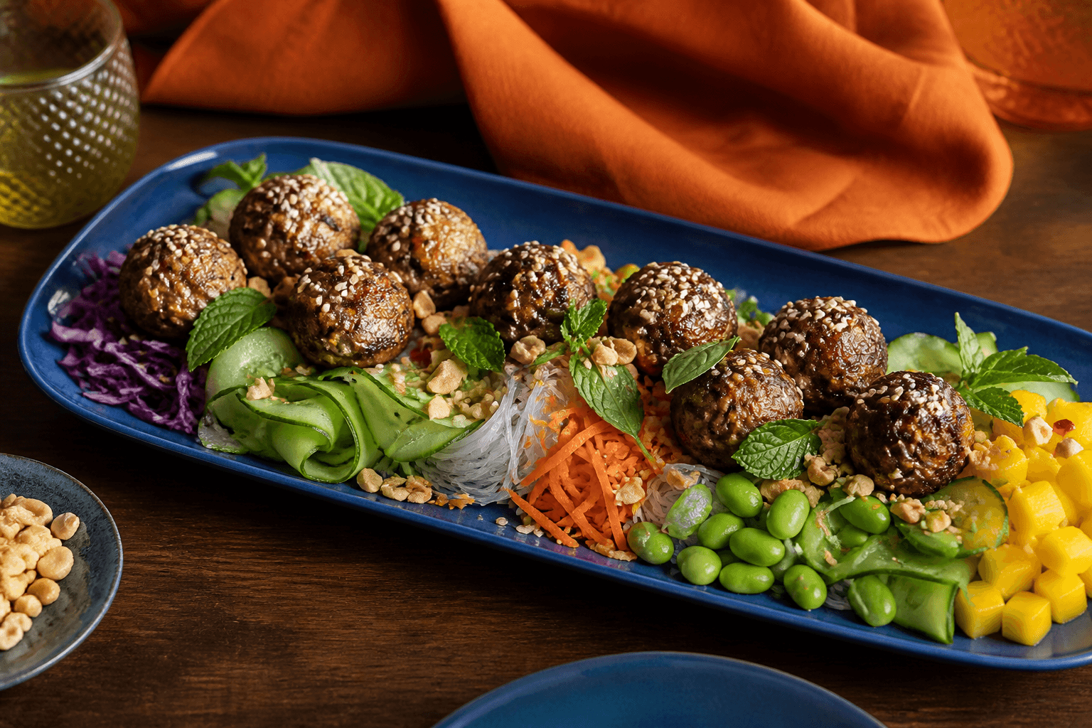 Langt koboltblåt fad med glasnudler, fuldt gennemstegte sesam-kyllingekødboller, rødkål, agurk, gulerod, edamame, mango, peanuts og urter