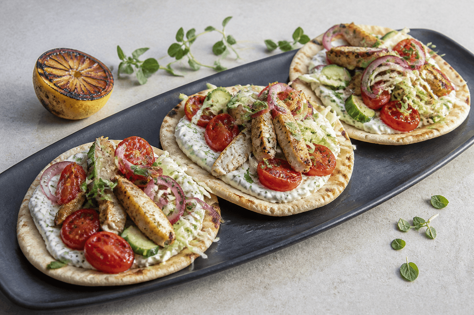 Langt sort stenfad med tre åbne gyrosfladbrød, fuldt tilberedt citron-oreganokylling, tomater, agurk, kål, syltet rødløg og mynte-tzatziki