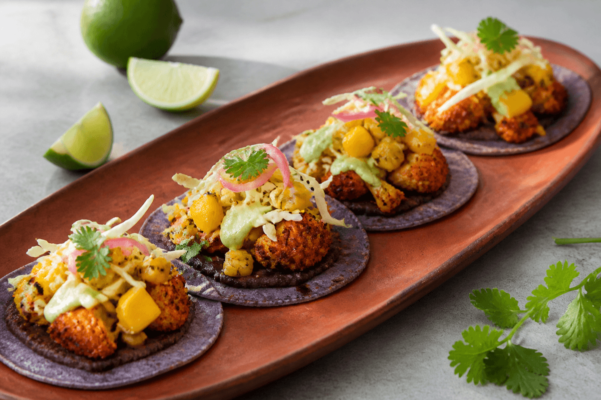 Langt terrakottafad med fire lilla majstacos, sorte bønner, ristet krydret blomkål, mango-majs-salsa, kål, syltet løg og avocadocreme