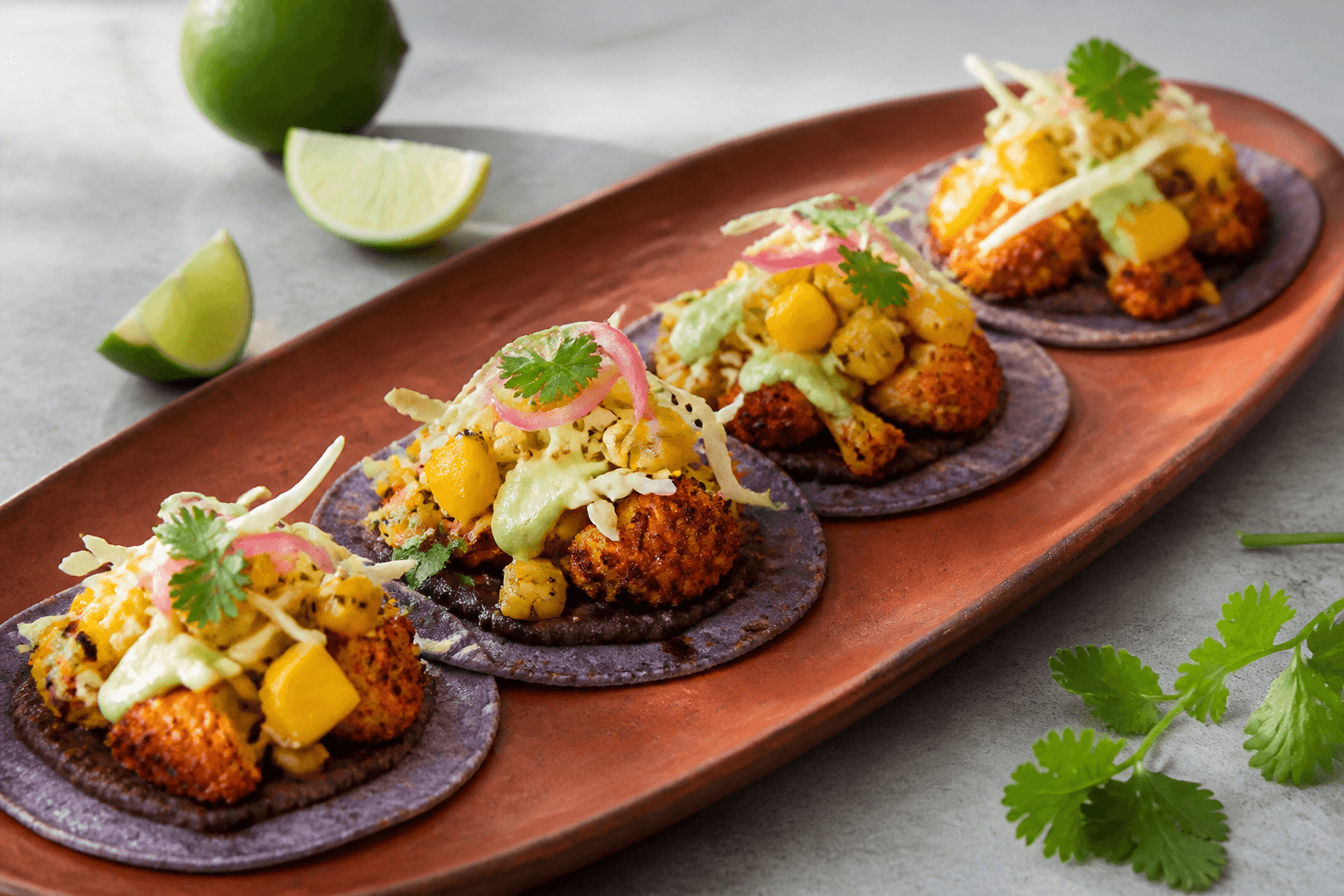 Langt terrakottafad med fire lilla majstacos, sorte bønner, ristet krydret blomkål, mango-majs-salsa, kål, syltet løg og avocadocreme
