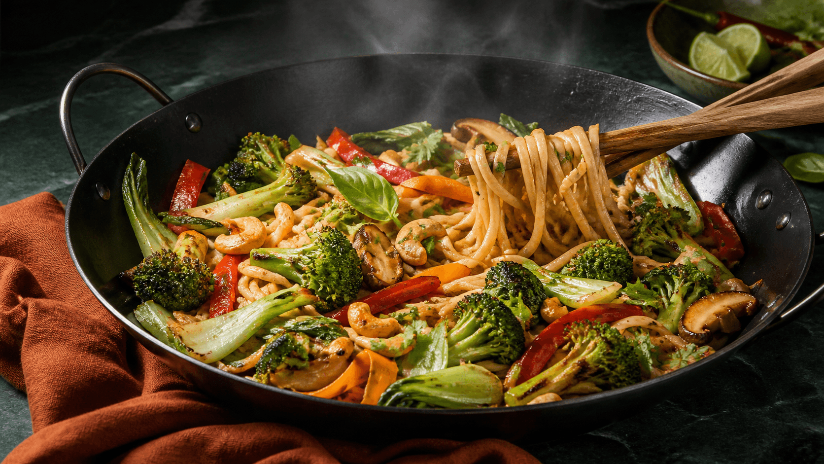 Sort stålwok med blanke kokos-limenudler, mørkt stegt broccoli, pak choi, rød peber, shiitake, gulerod, cashew, urter og løftet nudelbundt