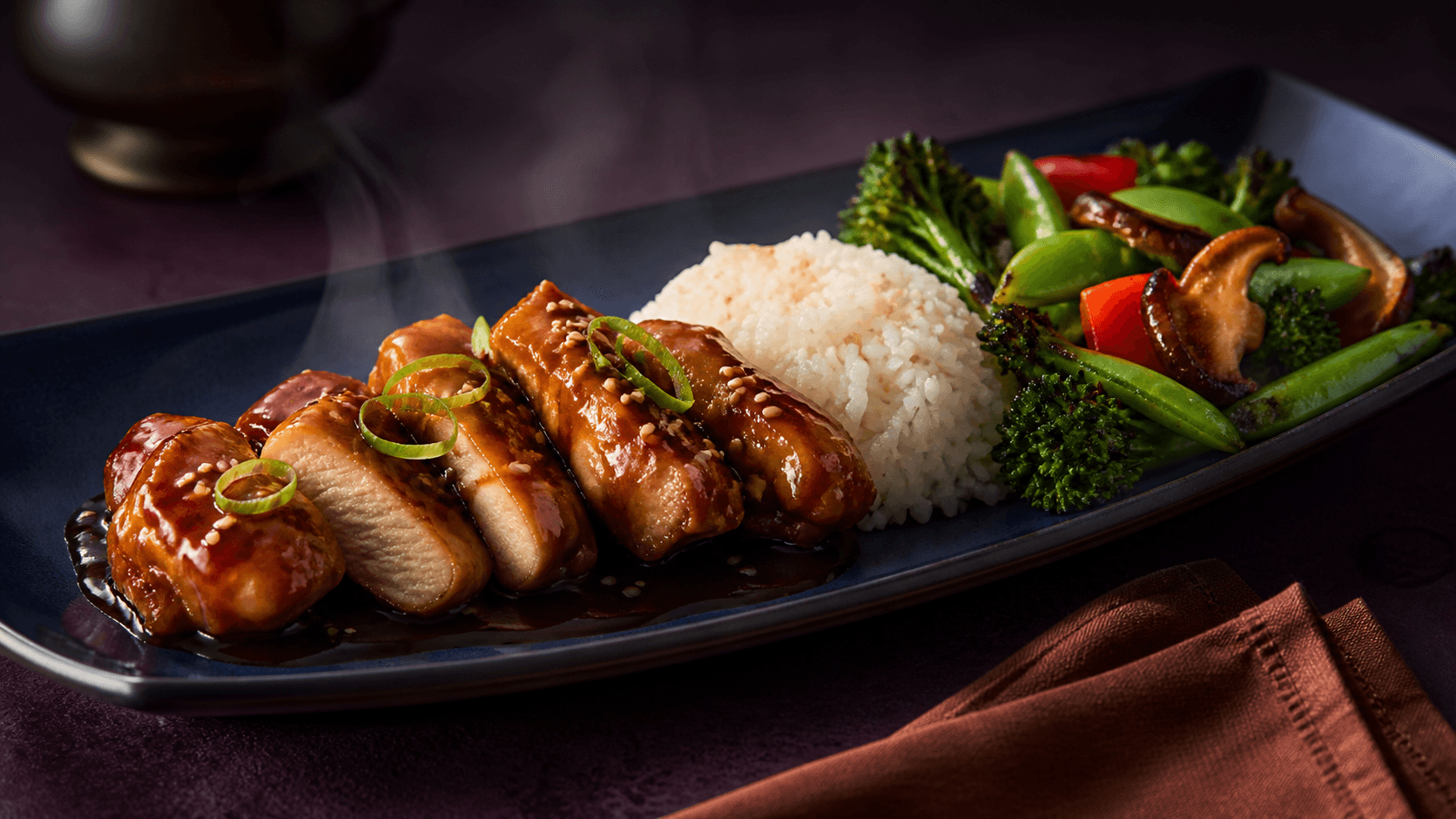Langt midnatsblåt fad med blank teriyakikylling gennemstegt til mindst 75 °C, åbent hvidt snit, jasminris, broccolini, sukkerærter, rød peber, shiitake, sesam og forårsløg