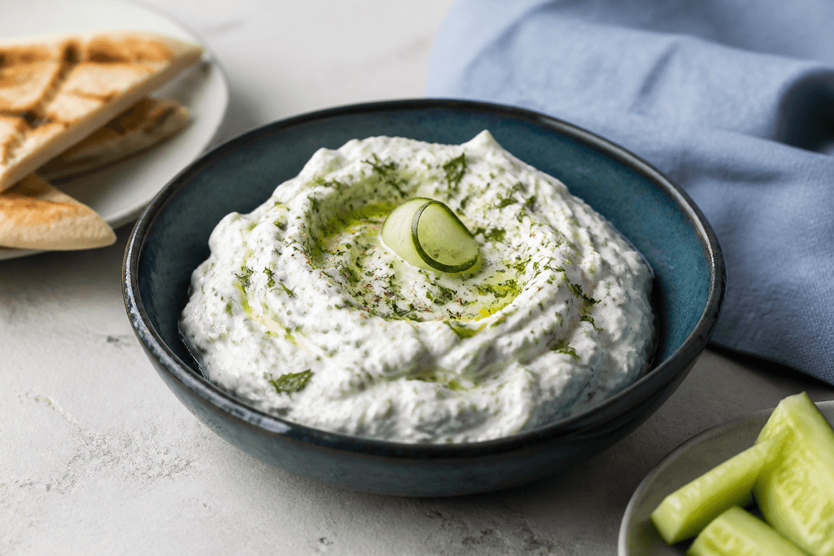 Mørk teal skål med tyk tzatziki, tydelige ske-riller, agurk, dild, mynte, citron, peber og olivenolie