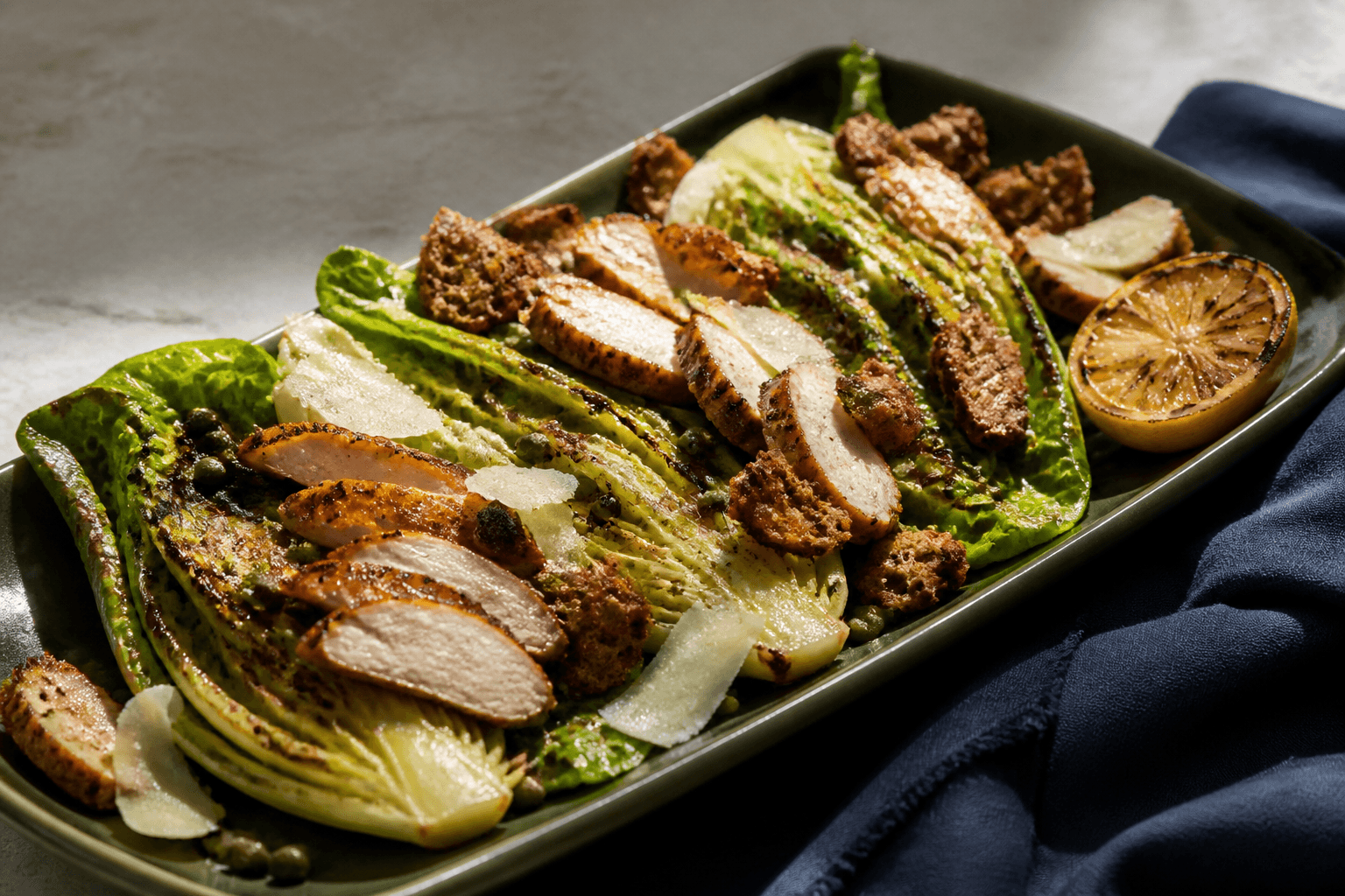 Langt skovgrønt fad med grillede romainehalvdele, helt gennemtilberedt kylling, parmesan, rugcrunch, kapers og grillet citron