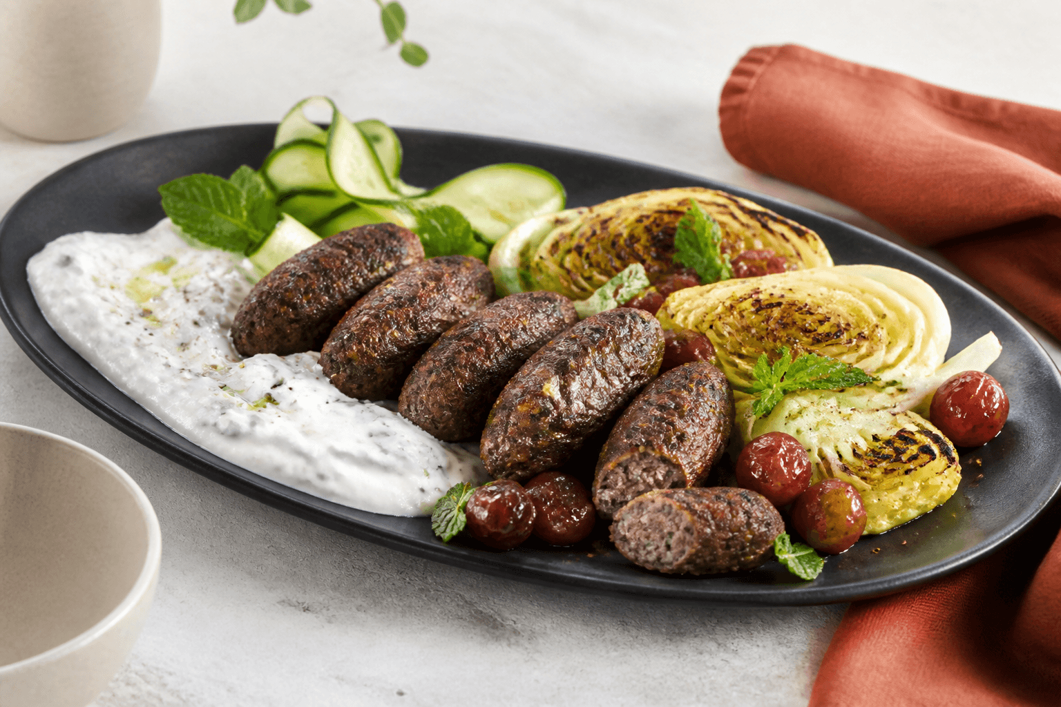 Lammekefta med grillede spidskålsbåde, feta-tzatziki, agurkebånd, bagte druer og mynte på langt sort fad