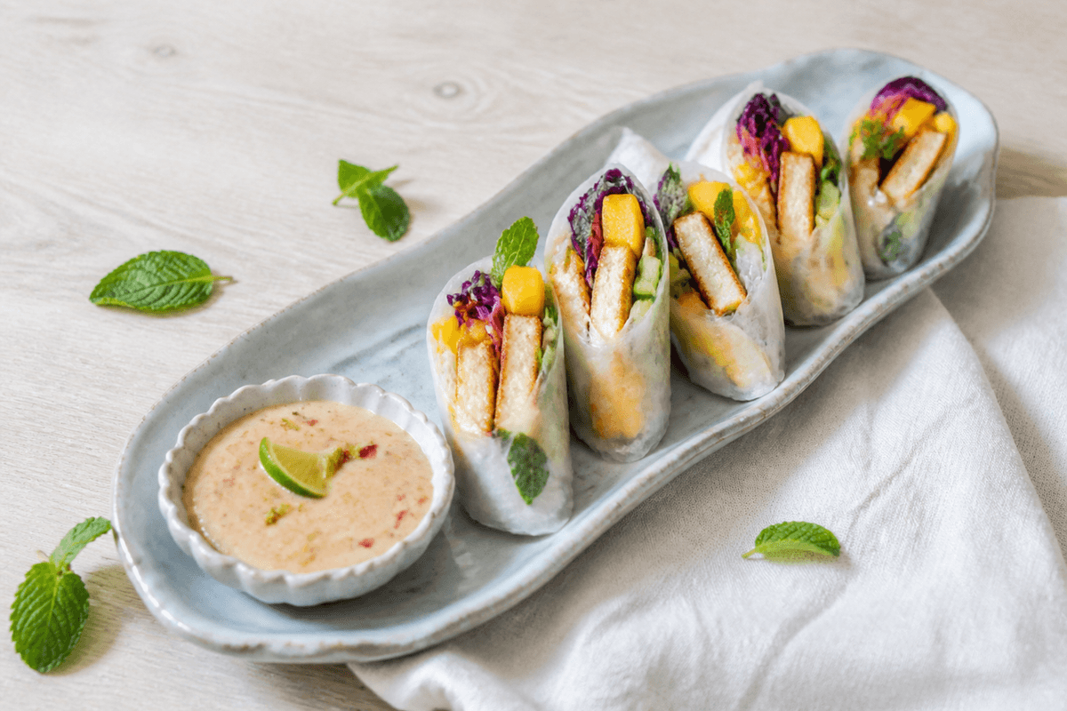 Lyst langt nordisk fad med halverede sommerruller fyldt med gylden halloumi, mango, lilla kål, agurk, mynte og risnudler samt cashew-limedip