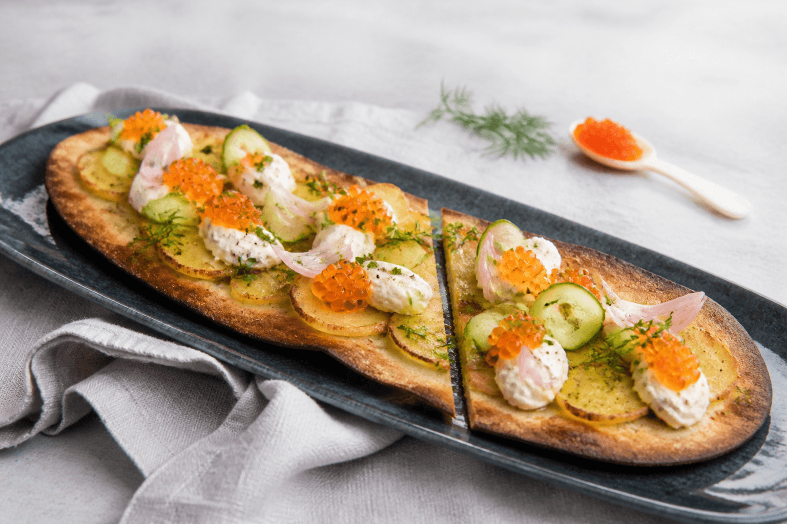 Langt tyndt rug-surdejsfladbrød med kartoffelskiver, pisket skyr-peberrod, stenbiderrogn, syltede skalotteløg, agurkebånd, purløg og dildolie på marineblåt fad
