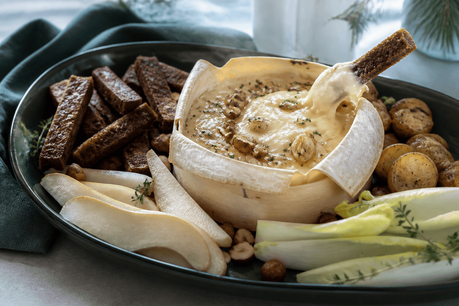Bagt Mont d'Or med timian, honning og hasselnød omgivet af rugbrødsstænger, kartofler, pære og julesalat på mørkegrønt fad