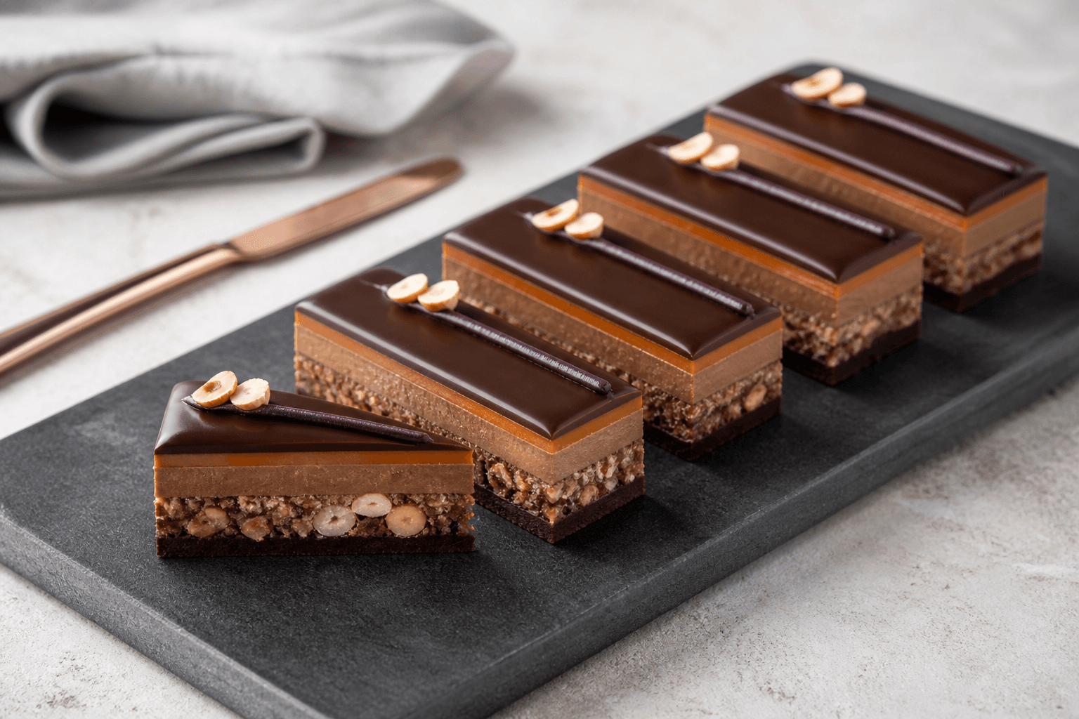 Lange lagdelte nougatsnitter med mørk chokolade, hasselnøddeknas, blød gianduja, tynd appelsinmarcipan og solbærlinje på mat koksgrå stenplade