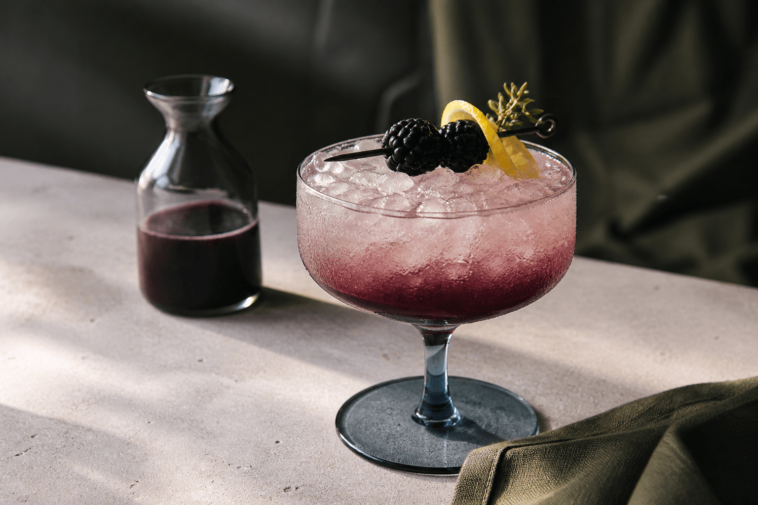 Nordisk bramblecocktail i lav røgblå coupe med knust is, brombær-solbær-ombré, citronskal og citrontimian ved lille karaffel