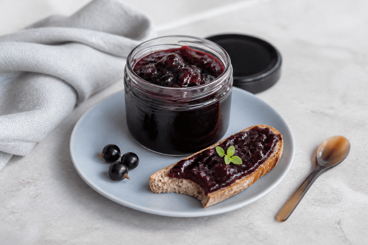 Mørk solbærmarmelade med ristet pære, citronverbena og sort peber i lille glas på lyseblå tallerken med ristet rugbrød