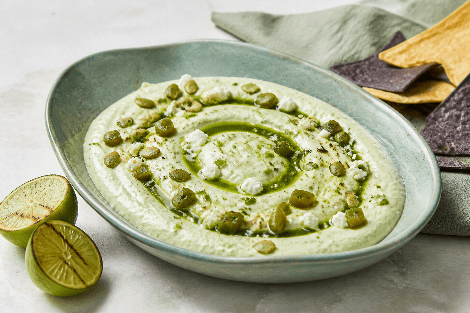 Grøn jalapeño-avocadodip med feta, korianderolie og ristede græskarkerner i et lavt celadongrønt fad med tortillaer og lime