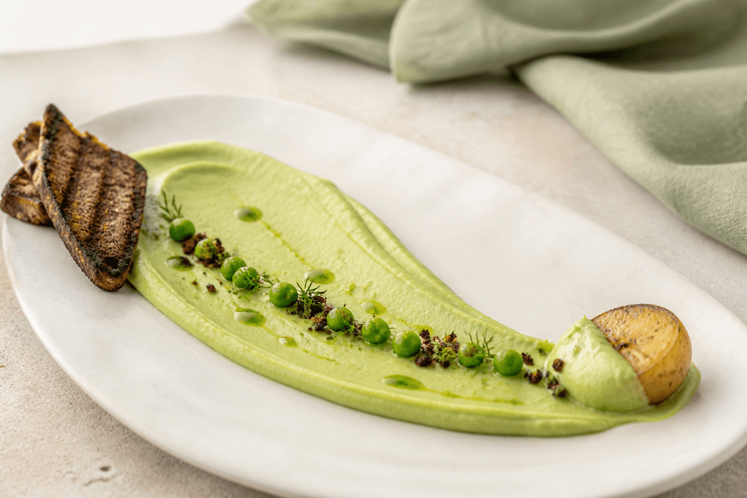 Silkeblød avocado-ærtecreme med dildolie, grønne ærter, rugknas og lime som langt grønt strøg på et lyst fad