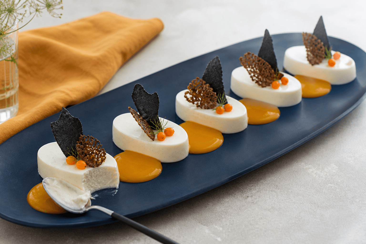 Fem kærnemælks-panna cotta med havtornscurd, lakridsmarengs, rugflorentin og friske havtorn på et langt mørkeblåt fad